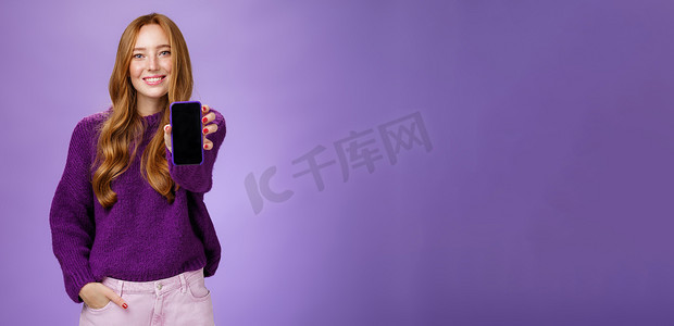女孩在相机前展示智能手机屏幕，询问朋友的意见，面带乐观和快乐的表情，手牵着口袋，在紫色背景下推销手机或应用程序