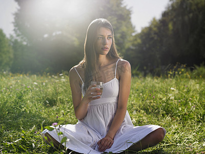 盘腿摄影照片_盘腿坐在草地上拿着水杯的年轻女人