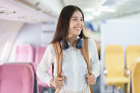 美女手提行李摄影照片_年轻的亚洲美女乘飞机旅行，乘客戴着耳机把手提行李放在飞机座位上方的储物柜里