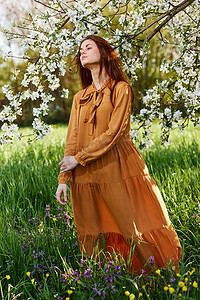 绿色树苗条摄影照片_一个苗条、甜美的女人穿着橙色长裙站在一棵开花的树旁高高的草丛中，看着别处，她的裙子在微风中展开