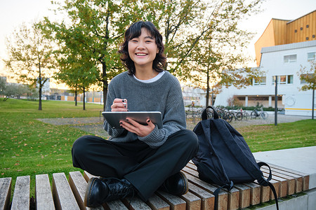 年轻的亚洲女孩，学生坐在长凳上，平面设计师用铅笔和数字平板电脑画画，在户外做作业