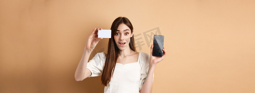 兴奋的女孩展示银行塑料信用卡和空手机屏幕，展示购物应用程序，站在米色背景中