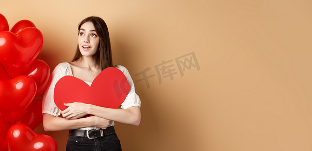 浪漫的女孩拥抱情人节大红色的心形剪纸，站在浪漫的气球旁边，向左看标志，梦幻般地凝视着促销，米色背景