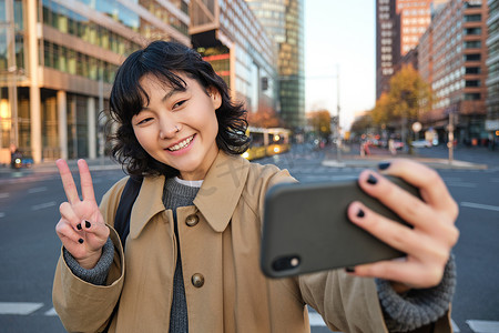 快乐的亚洲女孩，游客在市中心拍照，在智能手机相机上展示和平标志，在观光时自拍，面带微笑