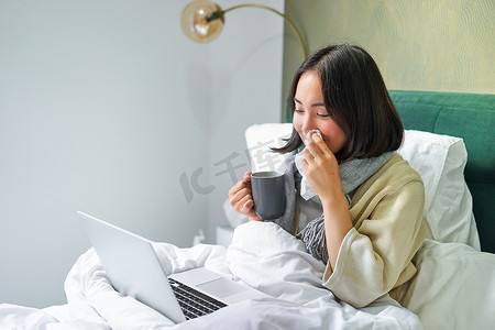 勤劳的亚洲女孩因感冒躺在床上，感染流感，但在家用笔记本电脑工作，一边生病一边学习