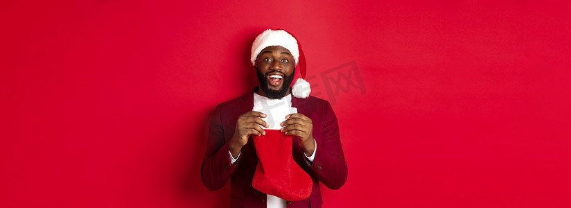 圣诞袜里有礼物摄影照片_兴奋的黑人打开圣诞袜，里面放着礼物和糖果，笑得很开心，站在红色背景下的圣诞帽里