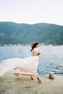 穿着随风飘扬的裙子的新娘拿着花束沿着码头走