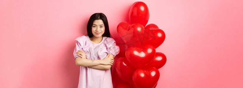 自信的少女双臂交叉在胸前微笑，穿着可爱的裙子，带着红心气球，站在粉红色背景中庆祝情人节