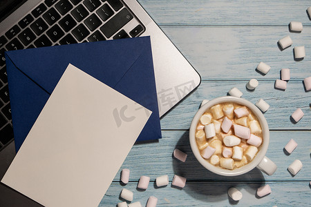 空的问候语或邀请卡模拟蓝色信封，白色咖啡杯和棉花糖笔记本电脑键盘放在蓝色木制桌子上。