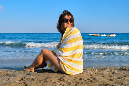 带沙滩巾的成熟美女坐在沙滩上，享受海上日落景观的女人