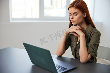 一位表情严肃、工作疲惫的女性坐在笔记本电脑前，双手交叉放在脸旁