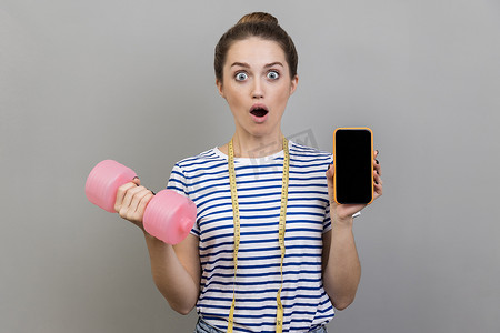 女人拿着粉红色的哑铃和带有空白屏幕的手机，用于宣传文字。