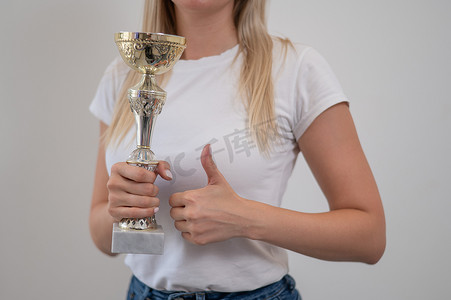 金发女人微笑着拿着金色的奖杯，在白墙上竖起大拇指。