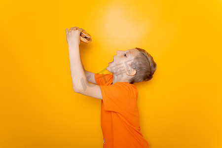 一个穿橙色T恤的男孩侧身站着，手里拿着一个他想吃的汉堡包