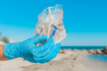 水在自然界的循环摄影照片_一个戴着蓝色家用防护手套的男人的手，在袋子里拿着一堆垃圾，贴着天空和海滩，特写
