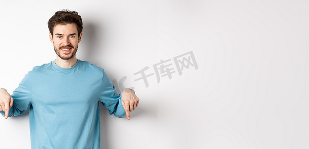微笑自信的男人手指向下，在白色背景上展示促销横幅或标志，站在休闲的蓝色运动衫中
