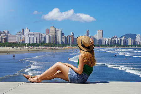 美丽的年轻女子拿着帽子坐在地上，大都市天际线，在模糊的背景下挥舞着海洋