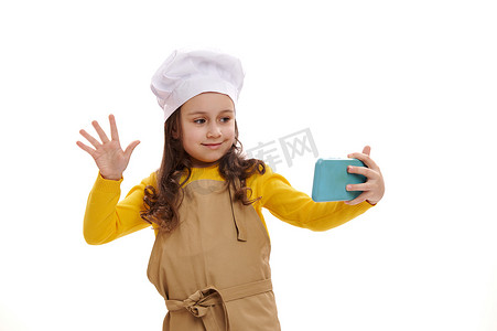 戴着白色厨师帽和厨房围裙的可爱小女孩，通过视频链接在手机上聊天，孤立的背景