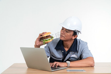 喜欢吃汉堡摄影照片_亚洲男工在家工作时微笑着拿着汉堡包在客厅里，下摆很开心，喜欢吃快餐。