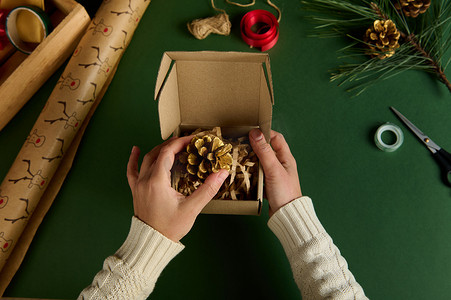 顶视图手将金色松果包装在绿色背景的工艺礼品盒中。