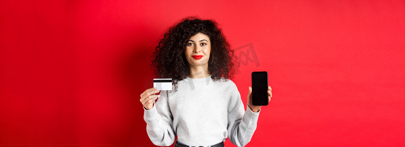 一头卷发的年轻现代女性展示塑料信用卡和手机屏幕，展示在线购物应用程序，站在红色背景中