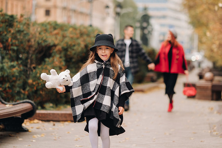 父母起跑摄影照片_一个小女孩和她的父母一起跑到秋天的城市。一个时尚的三口之家漫步在秋天的城市