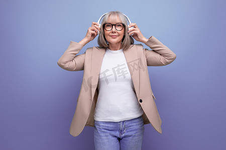 60 多岁的祖母穿着时尚的夹克，在明亮的背景下用大耳机听音乐，有复制空间