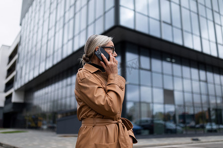 在办公中心玻璃立面的背景下，身穿秋装的商务高级成年女性用手机讲话，50 岁的欧洲女性，商业战略概念