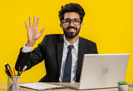 在笔记本电脑上工作的商人友好地对着镜头微笑，在办公室挥手打招呼