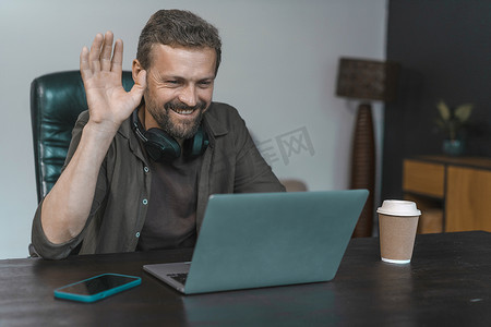 在现代阁楼办公桌上，白人男子通过笔记本电脑上的视频通话进行在线对话，办公桌上有电脑、手机和咖啡纸杯。