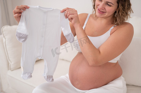 一位孕妇坐在白色沙发上，拿着待产婴儿的衣服。