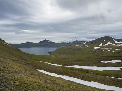 偏远的摄影照片_西峡湾美丽的 Hornbjarg 悬崖风景优美，冰岛偏远的 Hornstrandir 自然保护区，绿雪覆盖的山脉，蔚蓝的大海和多云的天空背景