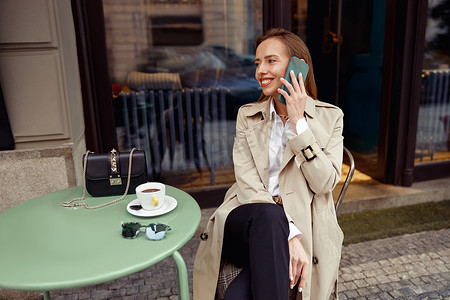 坐在咖啡馆露台上边喝咖啡边打电话的微笑时尚女性