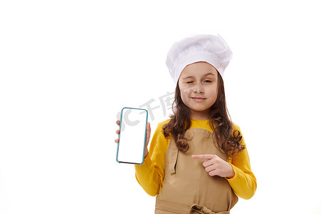 戴着厨师帽和围裙的小女孩，指着智能手机的白色空白数字屏幕，眨眼看着相机