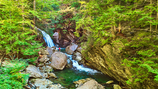 在郁郁葱葱的绿色森林的岩石峡谷中空中俯瞰河流，瀑布和孤独的徒步旅行者观察从巨石坠落