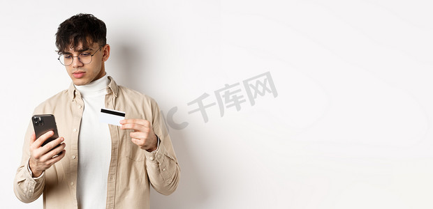 戴眼镜的帅气年轻人在电话上购物，在网上购物，拿着塑料信用卡和智能手机，站在白色背景上