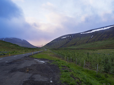 通往山脉的公路摄影照片_柏油路曲线穿过空旷的北部景观，绿草如茵的山峦和夕阳般的天空，通往冰岛西部高地的山脉