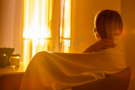 窗边浪漫摄影照片_浪漫的女孩裹着毯子，坐在客厅里，在窗边欣赏日落