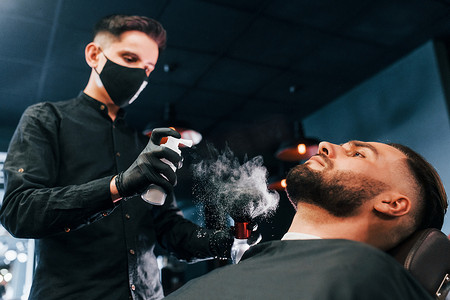 发型时髦的年轻人坐着，在理发店被戴黑色防护面具的男人剃胡子