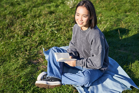 读书器摄影照片_亚洲女孩读书的画像，坐在公园的毯子上，绿草如茵，笑得开心
