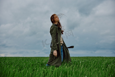 一个穿着外套的女人站在田野中间，在多云的天气里，在强风中侧身对着镜头