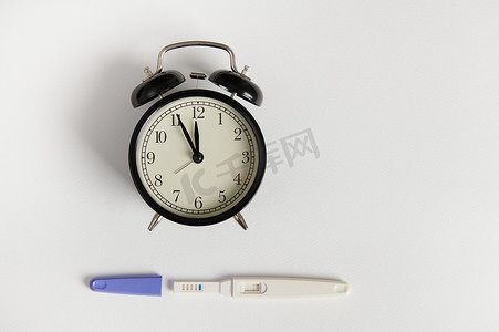平躺在白色背景上的闹钟和阳性妊娠试验套件。