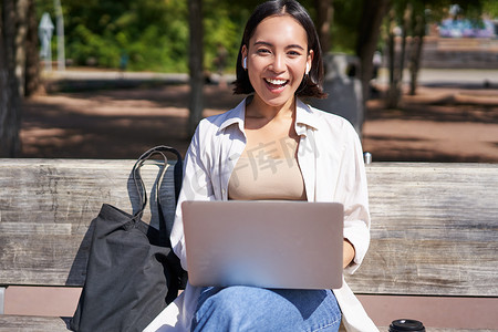 亚洲女性坐在公园长椅上，带着笔记本电脑，用无线耳机听音乐，做家庭作业，远程工作的肖像
