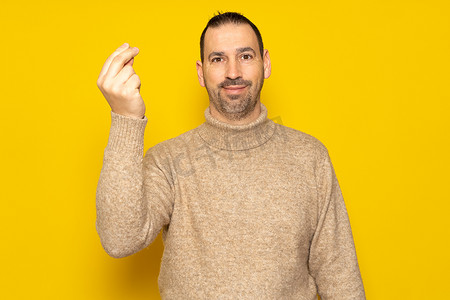 黄色手指手势摄影照片_留着胡子的西班牙裔男子身穿米色高领毛衣，一边对着镜头微笑，一边做着意大利妈妈咪呀的手势，在黄色背景中被隔离