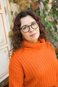 秋天的画像中，一位戴着时尚眼镜、身穿时尚橙色针织毛衣、背景为树叶的迷人年轻女子的画像。