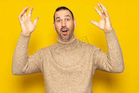 好看黄色摄影照片_40 多岁留着胡子的西班牙裔男子穿着米色高领毛衣，在收到一个巨大的惊喜后，精力充沛地举起双臂，在黄色工作室背景下被隔离。