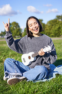 快乐的亚洲女孩在公园里弹尤克里里，展示摇滚乐、重金属喇叭标志和微笑，在户外玩得开心