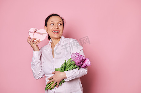 带着郁金香花束的迷人女人，摇着礼盒，试图猜测里面是什么，孤立的粉红色背景