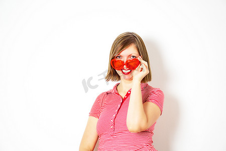 心形太阳镜摄影照片_戴着红色心形太阳镜的可爱微笑的年轻女子站着看着。