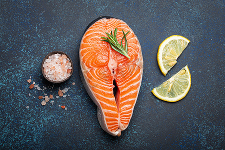 玫瑰盐摄影照片_深蓝色质朴石背景上带迷迭香的新鲜生鲑鱼片牛排，玫瑰盐和柠檬顶视图，健康均衡的饮食和营养